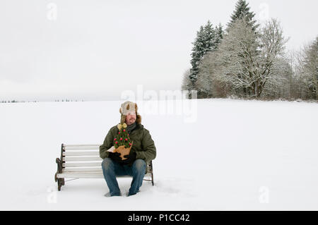 Ein Mann sitzt auf einer Bank im Schnee, mit Schnee bedeckten Bäume hinter ihm. Stockfoto