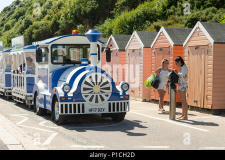 Bournemouth, Dorset, England, Großbritannien, heißes und sonniges Wetter im Juni. Blauer Landzug auf der Promenade vorbei an orangefarbenen Strandhütten und zwei jungen Frauen in Sommershorts. Stockfoto