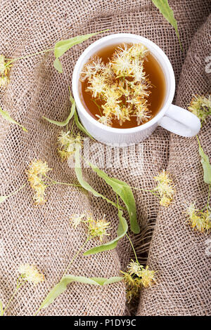 Tee mit einem Linden auf einem hölzernen Tisch, eine Tasse steht auf einer Leinwand Serviette. Um die Schale linden Blüten verstreut Stockfoto