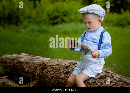 Ein litttle zwei Jahre alte Junge sitzt auf einem Baum und liest ein Buch. hildren Tag. Stockfoto