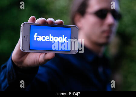 Nahaufnahme des ernsten jungen Mann mit Sonnenbrille, weiß iPhone mit Facebook Logo auf dem Bildschirm Stockfoto