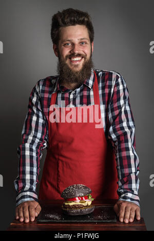 Männer kochen in rot Schürze in der Nähe von Frisch selbst gemacht schwarze Burger und lächelnd Stockfoto