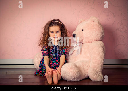 Kleines Mädchen spielt großen weichen Bären in einem rosa Zimmer, in einem schönen Kleid Stockfoto