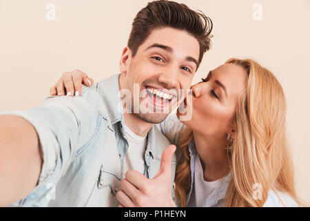 Portrait von glücklichen Menschen, die selfie Foto und deutete mit dem Daumen nach oben an der Kamera, während adorable Frau Küssen seine Wange über Gelb Hintergrund isoliert Stockfoto