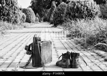 Mit leichtem Gepäck reisen! Abgenutzte Ankle Boots neben einem vintage Karton Koffer, ein Film Kamera in seine offene Ledertasche und einem brick road. Ein S/W Foto Effekt. Stockfoto