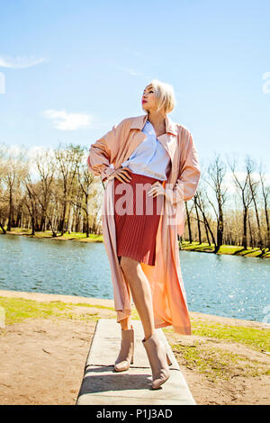 Perfekte Frau tragen Rosa seidige Regenmantel, Rock, Bluse und Ankle Boots, Street Fashion Portrait im Freien Stockfoto