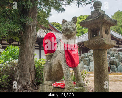 Shinto Kitsune fox Wächter mit roten Latz und gestrickte Schwanz, Taisanji Tempel 52, Shikoku 88 Tempel Wallfahrt, Ehime, Japan. Stockfoto