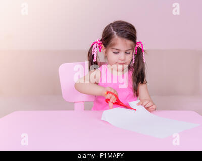 Portrait von Cute girl schneidet Papier mit einem roten Schere beim Sitzen im pink Tabelle. Selektiver Fokus und kleine Tiefenschärfe. Stockfoto