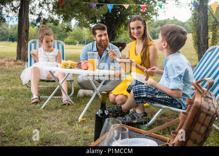 Engagierte junge Eltern von zwei Kindern hören auf Ihre lustigen Sohn sprechen beim zusammen essen während der Familie Picknick in einem Sommertag Stockfoto