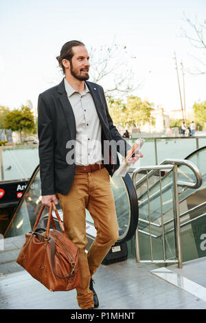 Volle Länge Bild der erfolgreiche Geschäftsmann im stilvollen Gesellschaftsmode, die Rolltreppe im Stadtzentrum mit Leder männliche Tasche und Zeitung in der Hand Stockfoto