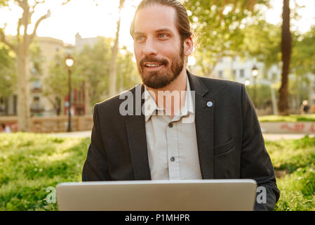 Bild Nahaufnahme von sachlich gut aussehender Mann mit gebunden Haar arbeiten an Silber Laptop während im City Park während der sonnigen Tag ruhen Stockfoto