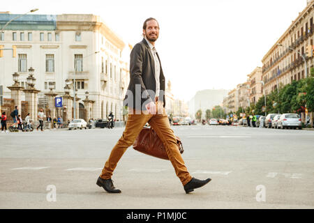 Portrait von eleganten Geschäftsmann in Jacke Lachen beim Überqueren der Straße in Megapolis mit Leder männliche Tasche in der Hand Stockfoto
