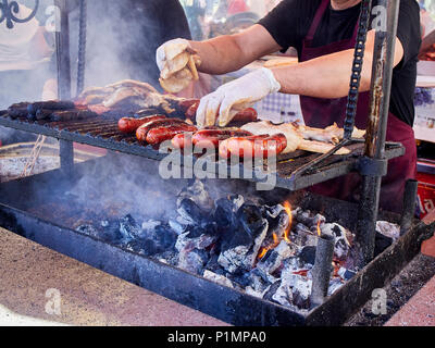 Koch, Würstchen, Speck und andere Fleisch auf einem Holzkohle Grill in einem Stall eines Street Food Fair. Stockfoto