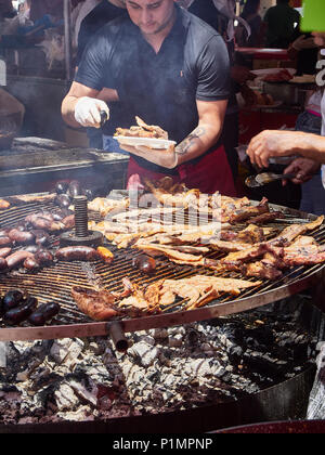 Hispanic Koch, Würstchen, Schweinerippchen und andere Fleischsorten Schnitte auf Holzkohle Grill in einem Stall eines Street Food Fair. Stockfoto