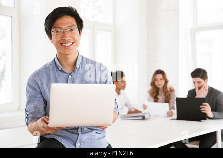Glückliche junge asiatische Mann mit Laptop und lehnte sich auf eine Tabelle mit sprechenden Kolleginnen und Kollegen auf einem Hintergrund Stockfoto