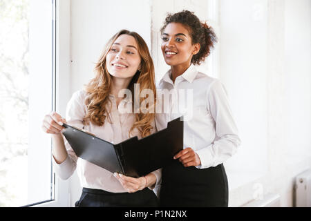 Zwei lächelnde business Frauen sitzen im Innenbereich mit einer offenen Ordner und Wegsehen Stockfoto