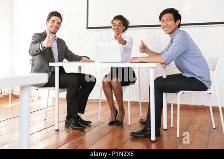 Gruppe von lächelnden Jungen multiethnischen Geschäftsleute sitzen am Schreibtisch und geben Daumen bis Stockfoto