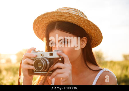 Closeup Bild der schönen jungen Frau mit langen dunklen Haaren tragen Strohhut Fotografieren auf retro Kamera, während in der sonnendurchfluteten Natur Ruhe Stockfoto