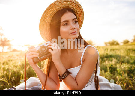 Bild der Landschaft Mädchen 20 s mit langen dunklen Haare trug Stroh Hut und weißen Kleid Holding retro Kamera während der Sitzung auf Gras im Park bei Sonnenaufgang Stockfoto