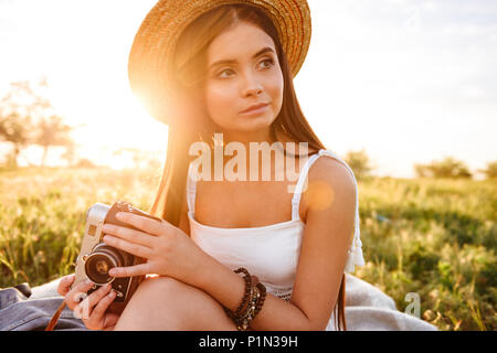 Bild der Landschaft Mädchen 20 s mit langen dunklen Haare trug Stroh Hut zur Seite suchen, und halten retro Kamera während der Sitzung auf Gras bei Sonnenaufgang Stockfoto