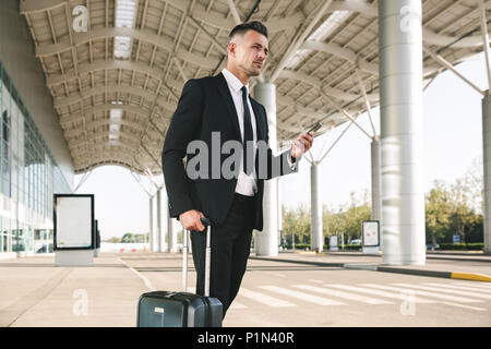 Zuversichtlich Geschäftsmann in Anzug gekleidet, gehen mit einem Koffer außerhalb Airport Terminal und Holding Handy Stockfoto