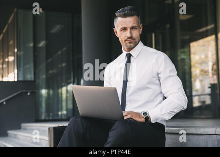 Porträt eines erfolgreichen jungen Geschäftsmann in formelle Kleidung außerhalb eines Gebäude aus Glas sitzen und Arbeiten am Laptop gekleidet Stockfoto
