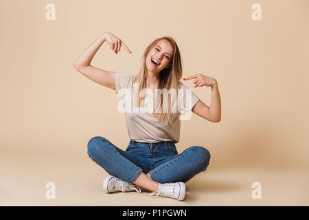 Foto der schönen Frau zeigt mit dem Finger auf sich selbst beim Sitzen auf dem Boden im Lotus in Freizeitkleidung pose über beigen Hintergrund isoliert Stockfoto