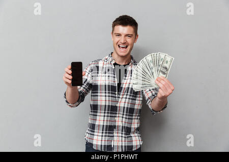 Porträt von einem Aufgeregten jungen Mann mit Geld Banknoten und leerer Bildschirm Mobiltelefon über grauer Hintergrund Stockfoto