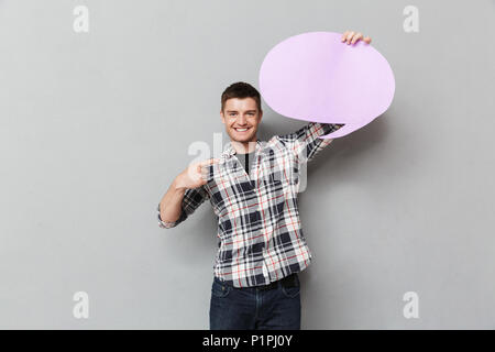 Porträt eines lächelnden jungen Mann in Plaid Shirt Zeigefinger auf Leerer Bildschirm Sprechblase über grauer Hintergrund Stockfoto