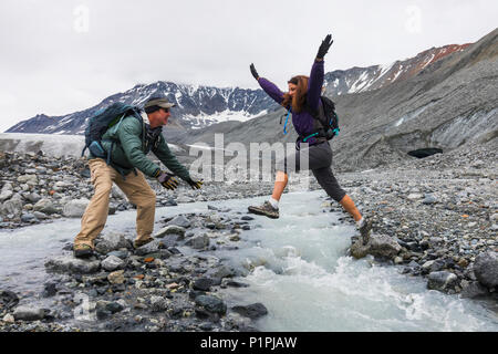 Eine Frau springt zu ihrem Ehemann in einem eisigen Strom beim Wandern in der Nähe von Gulkana Glacier; Alaska, Vereinigte Staaten von Amerika Stockfoto