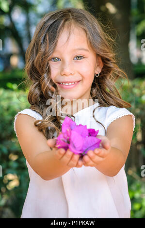 Porträt einer Kaukasischen fünf Jahre alten Mädchen lächelnd und mit violetten Blüten in einem Park als Geschenk Stockfoto