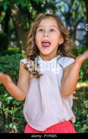 Cute fünf Jahre alten Mädchen mit blauen Augen und blonden Haaren Glück zeigen und überrascht auf Ihrem Gesicht mit einem Park als Hintergrund Stockfoto