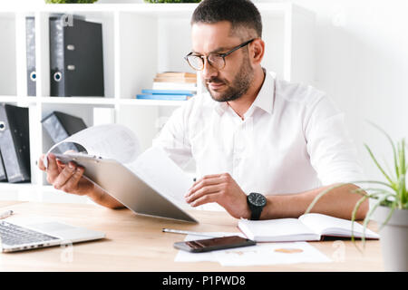 Bild von Konzentriertem unrasierte Geschäftsmann 30 s in Weiß shirt Holding und Zwischenablage mit Papier Dokumente während der Arbeit im Büro Stockfoto