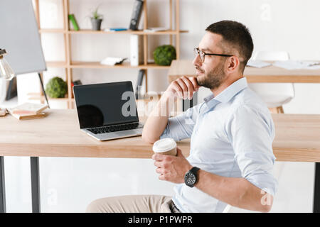 Profil Bild der sorgfältigen Büro Mann 30 s in Weiß Shirt sitzen am Tisch und trinken Kaffee zum Mitnehmen während der Arbeit am Laptop im Business Center Stockfoto