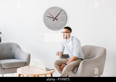 Bild des fleißigen Büro Mann 30 s im weißen Hemd sitzt im Sessel und trinken Kaffee zum Mitnehmen während der Arbeit am Laptop im Business Center Stockfoto