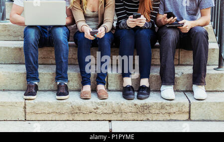 Vier Studenten in einer Reihe sitzen auf einem Schritt mit ihrer Technologie auf dem Campus der Universität, nur mit einem Blick auf ihre Füße und Beine Stockfoto
