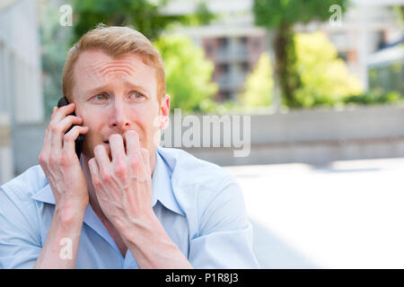 Closeup Portrait, besorgt, junger Mann im blauen Hemd am Telefon sprechen zu jemand, düster, isoliert im Freien außerhalb Hintergrund Stockfoto