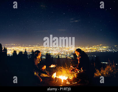 Glückliches Paar Wanderer wärmen Sie die Hände in der Nähe von Lagerfeuer unter Sternenhimmel mit Sterne und die Lichter der Stadt im Hintergrund Stockfoto