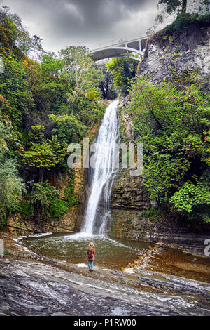 Touristische Frau in Hat in der Nähe des Wasserfalls im Botanischen Garten mit Bäume im Herbst an bedeckt bewölkten Himmel in Tiflis, Georgien Stockfoto