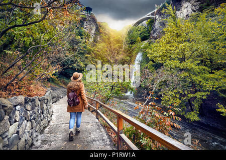 Touristische Frau in Hut gehen zu Wasserfall im Botanischen Garten mit Bäume im Herbst an bedeckt bewölkten Himmel in Tiflis, Georgien Stockfoto