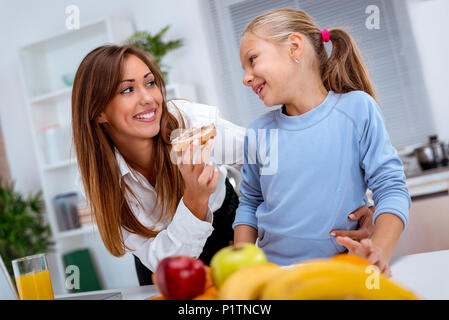 Junge schöne Mutter und ihre süße Tochter frühstücken in der Küche. Mutter ist die Tochter mit Scheibe Brot mit Pastete. Stockfoto