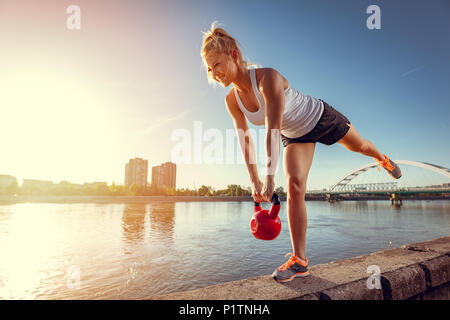 Junge fitness Frau tun Workout mit kettlebell auf der Wand, die mit dem Fluss in einem Sonnenuntergang. Stockfoto