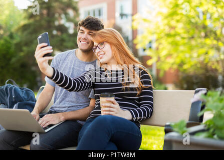 Ein junger Mann und eine junge Frau zusammen auf einer Bank auf dem Campus der Universität, die ein Selbst sitzen - Portrait auf einem Smartphone beim Trinken von Kaffee Stockfoto