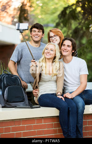 Eine Gruppe von vier Freunden auf einer Bank sitzen, ein Selbstporträt mit einem Smartphone mit einem selfie Stick auf einem Campus der Universität Stockfoto