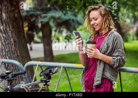 Porträt einer schönen jungen Frau Studentin lehnte sich auf einem Fahrradträger auf dem Campus und lesen Texte auf Ihrem Smartphone Stockfoto