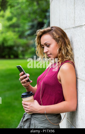 Eine schöne junge Frau Studentin gegen eine Wand auf dem Campus schiefen und lesen Texte auf Ihrem smart phone; Edmonton, Alberta, Kanada Stockfoto