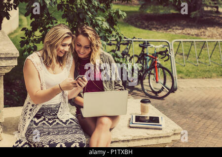 Zwei weibliche Studenten zusammen zu sitzen und über ihr Smartphone und Laptop sitzend auf die Schritte eines Campus Gebäude Stockfoto