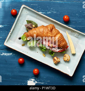 Lecker Sandwich aus Croissants gefüllt mit Gemüse, Salat, Pilzen und Sauce liegen auf flachen, rechteckigen Platte stehend auf Blau alte hölzerne Stockfoto