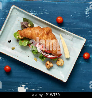 Lecker Sandwich aus Croissants gefüllt mit Gemüse, Salat, Pilzen und Sauce liegen auf flachen, rechteckigen Platte stehend auf Blau alte hölzerne Stockfoto