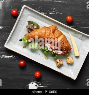 Lecker Sandwich aus Croissants gefüllt mit Gemüse, Salat, Pilzen und Sauce liegen auf flachen, rechteckigen Platte stehend auf Blau alte hölzerne Stockfoto